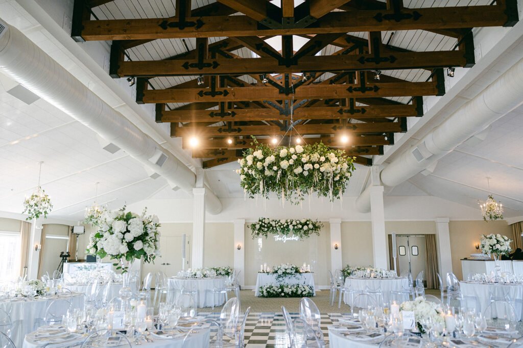A photo of The Vineyard Pavilion decorated for a wedding with florals all around and hanging from the chandelier. White round tables with chairs set up for dinner, with a dance floor in the middle.